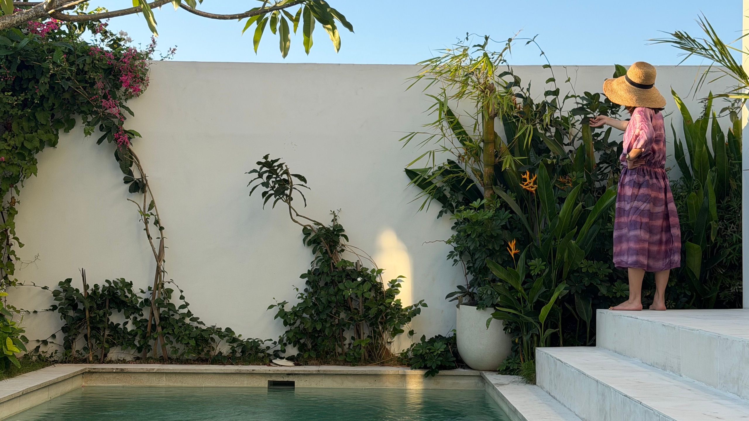 A woman in a straw hat on the marble pool steps beside the white wall and bougainvillea, in late afternoon light.