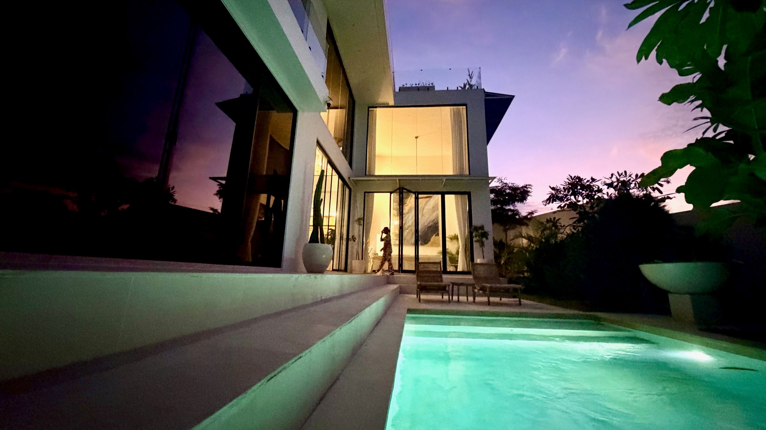 The white villa lit from within at twilight, glass glowing warm above the plunge pool, against a violet Bukit sky.