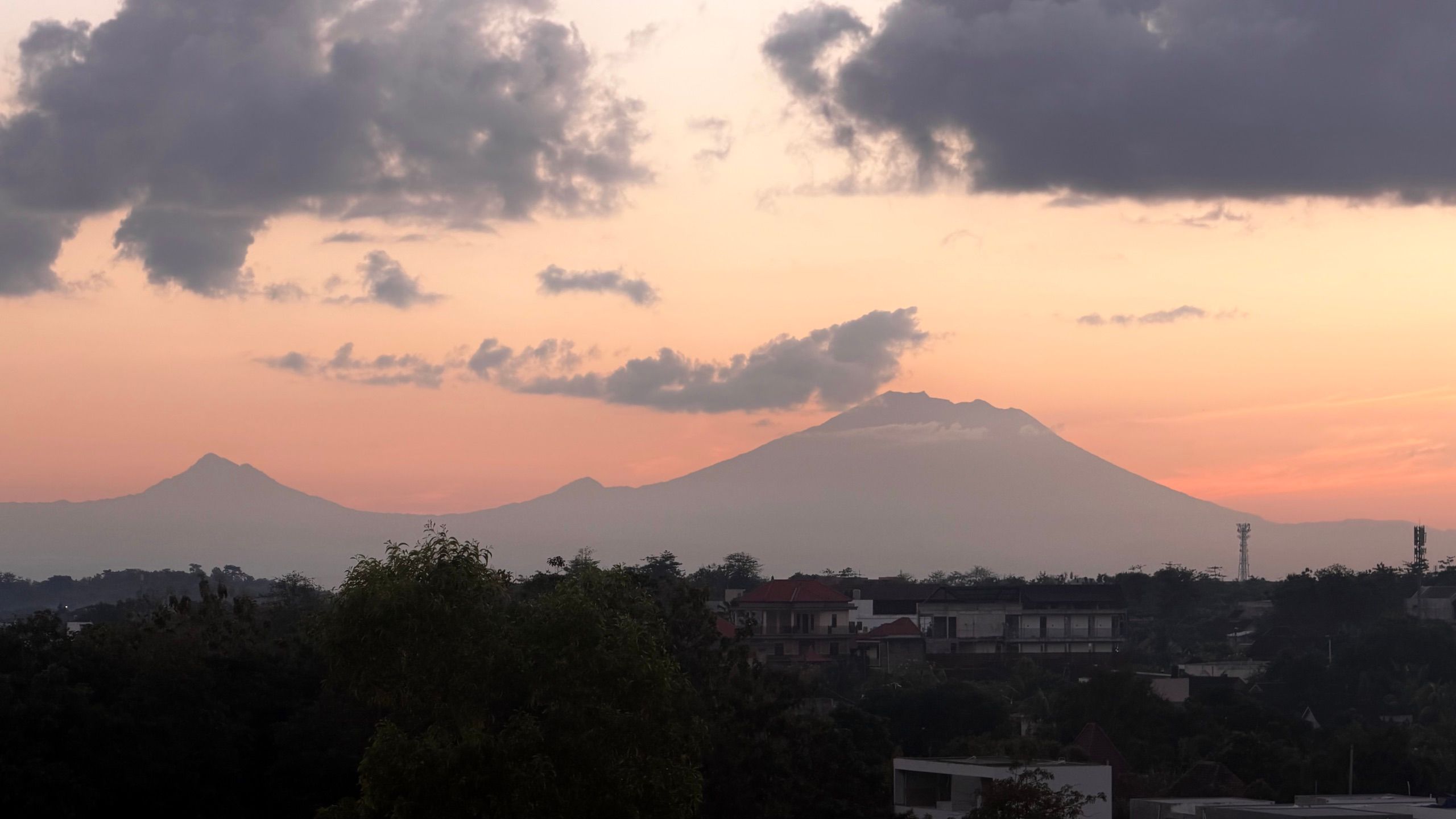 Mount Agung in silhouette under an orange dusk sky, seen across the rooftops of the Bukit.